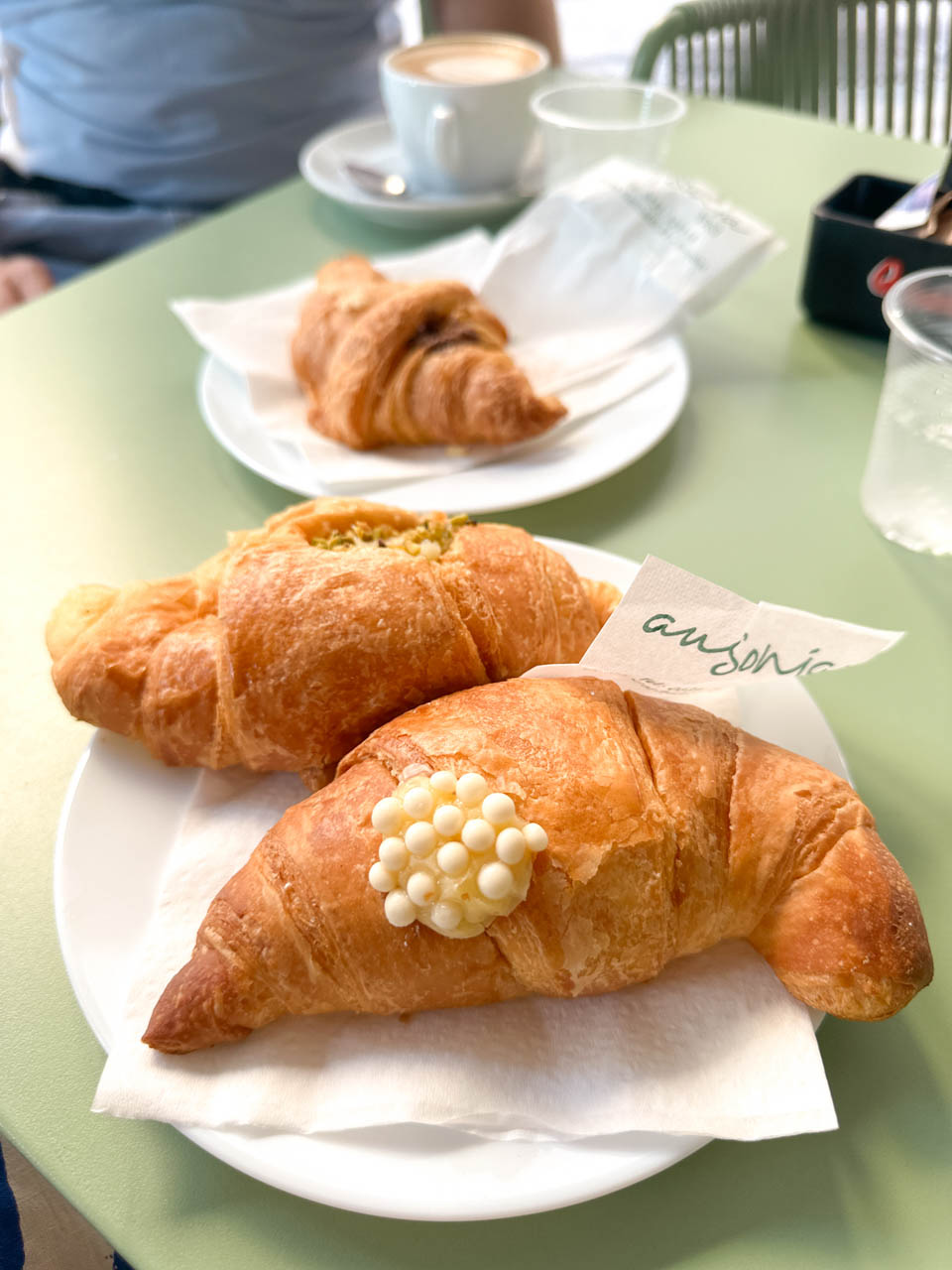Two filled croissants on a plate at a café table, with cappuccino and another croissant in the background