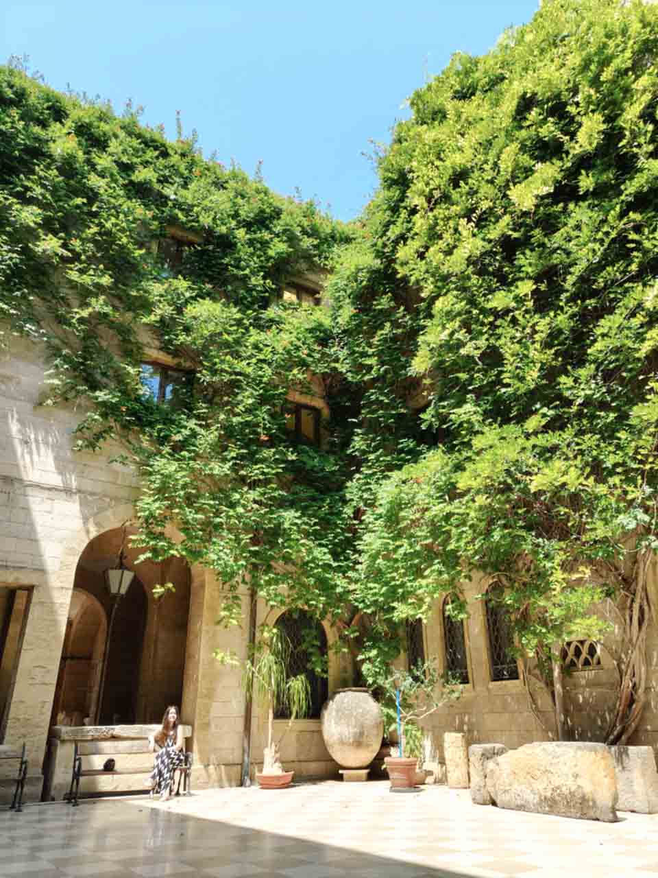 Courtyard of the Museo Archeologico Francesco Ribezzo in Brindisi, with ivy-covered walls, stone arches, potted plants, and a woman sitting on a bench in the shade