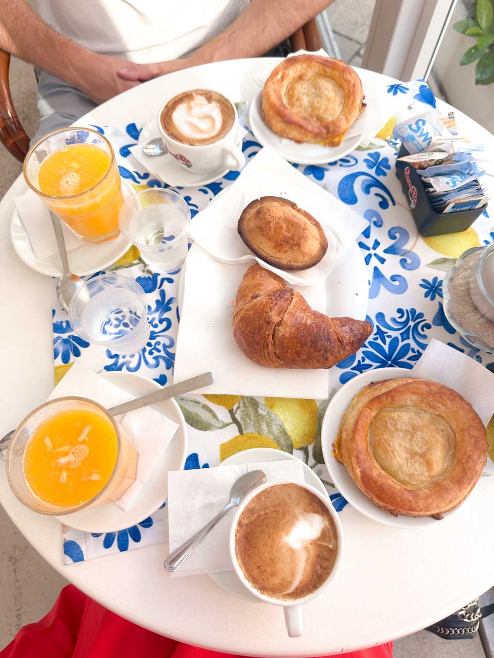 Breakfast table with a pasticciotti, a croissant, two rustico pastries, fresh orange juice, and cappuccinos on a lemon-print tablecloth