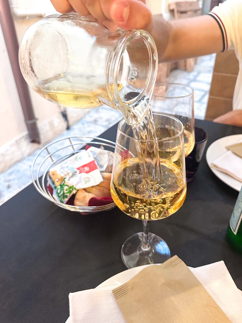 White wine being poured into a glass at an outdoor restaurant table with snacks in the background