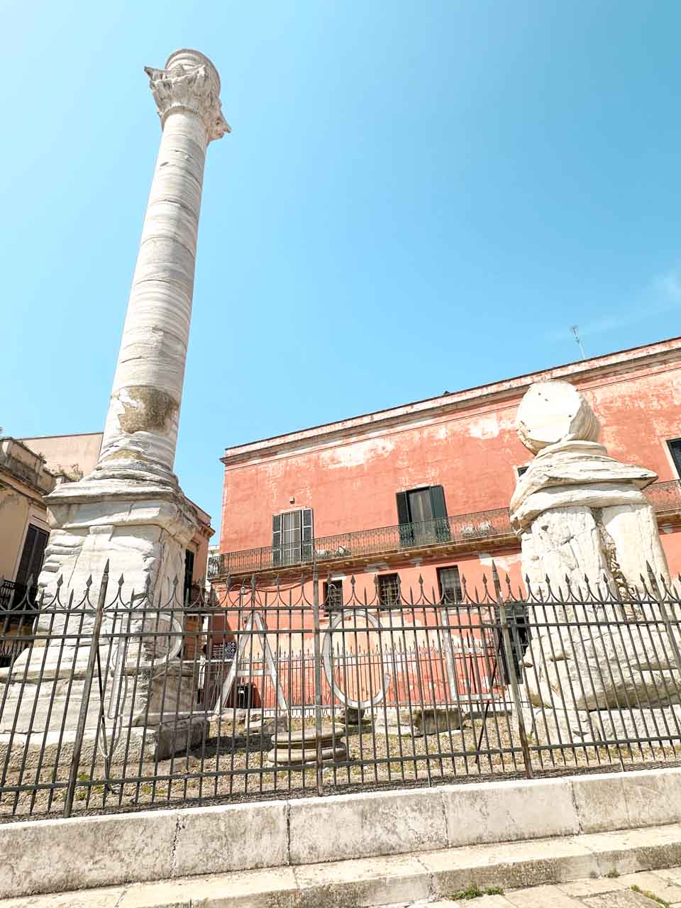 Close-up of the Roman columns in Brindisi, with a pink building and black iron fence behind