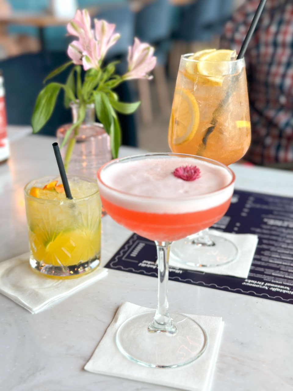 Three colourful cocktails on a table with a menu and a pink flower vase at the Vidokówka restaurant in Gdańsk
