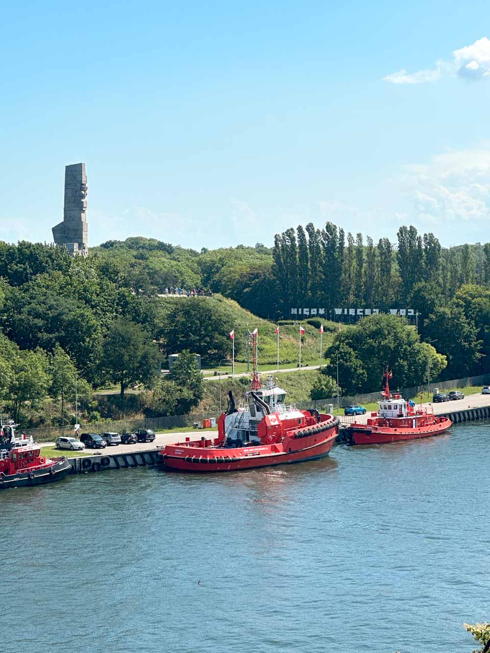 Red tugboats docked by Westerplatte, with the towering WWII Monument visible on the green hill behind