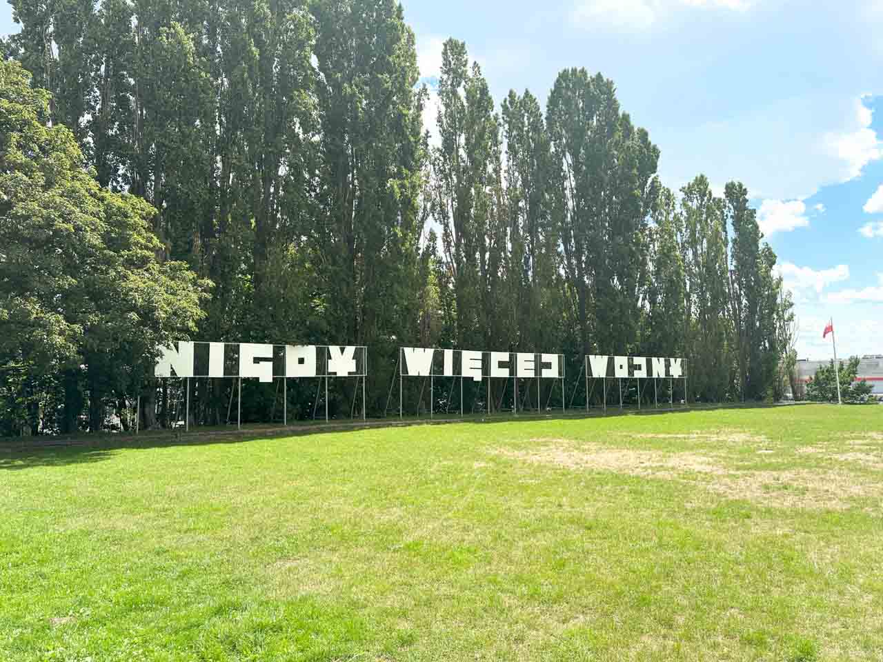 Large white “Nigdy więcej wojny” sign on Westerplatte, framed by tall trees and a grassy lawn