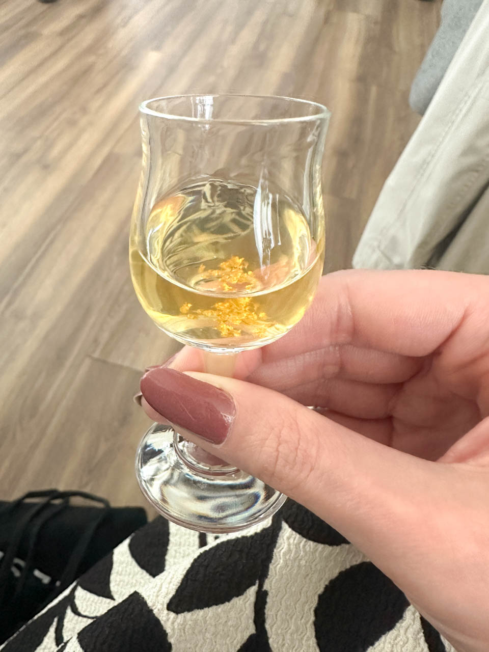 Hand holding a glass of Goldwasser liqueur with floating gold flakes over a wooden floor