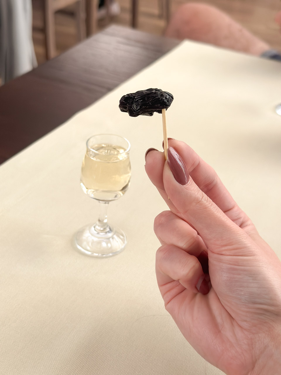 Close-up of a dried plum on a cocktail stick, held above a glass of Machandel liqueur