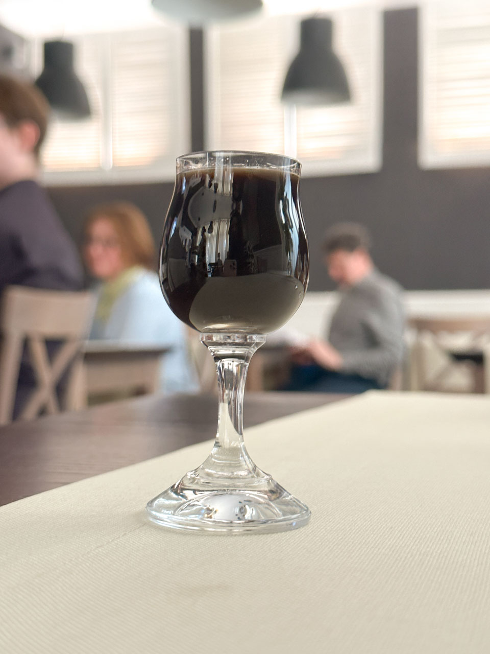 Glass of dark Kurfürsten liqueur on a cream tablecloth, with people blurred in the background