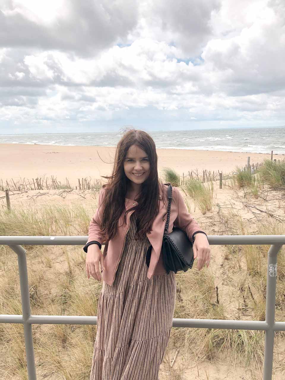 A woman smiling by the beach in a long striped dress and pink jacket, with the sea and sand dunes behind her