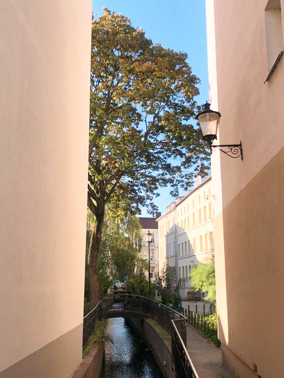 A small canal with a tree-lined pedestrian bridge between pastel buildings in Gdańsk