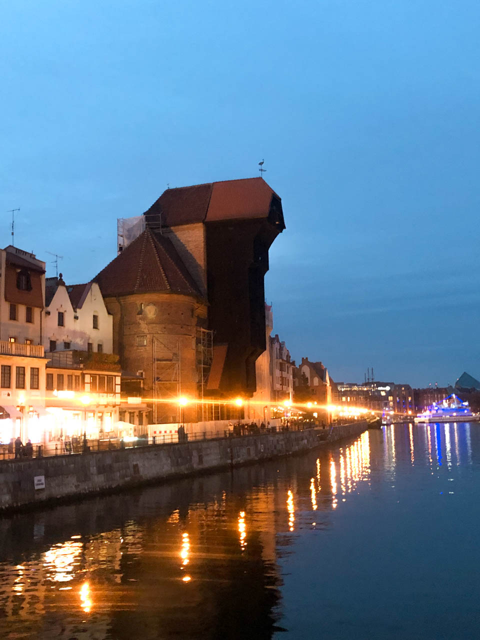 The medieval Gdańsk Crane by the Motława River at dusk, lit by streetlights reflecting in the water