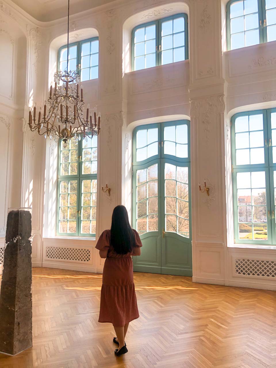 A woman in a pink dress walking through a grand room with pastel green windows and elegant chandeliers at the Abbots' Palace in Oliwa