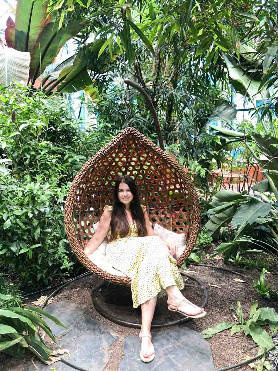 A woman in a yellow dress sitting on a woven hanging chair in a lush indoor tropical garden