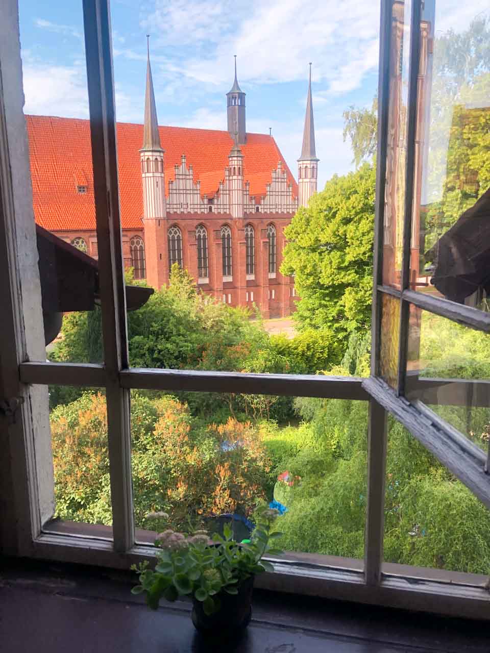 View of a red brick cathedral with spires seen through an open wooden window above a garden