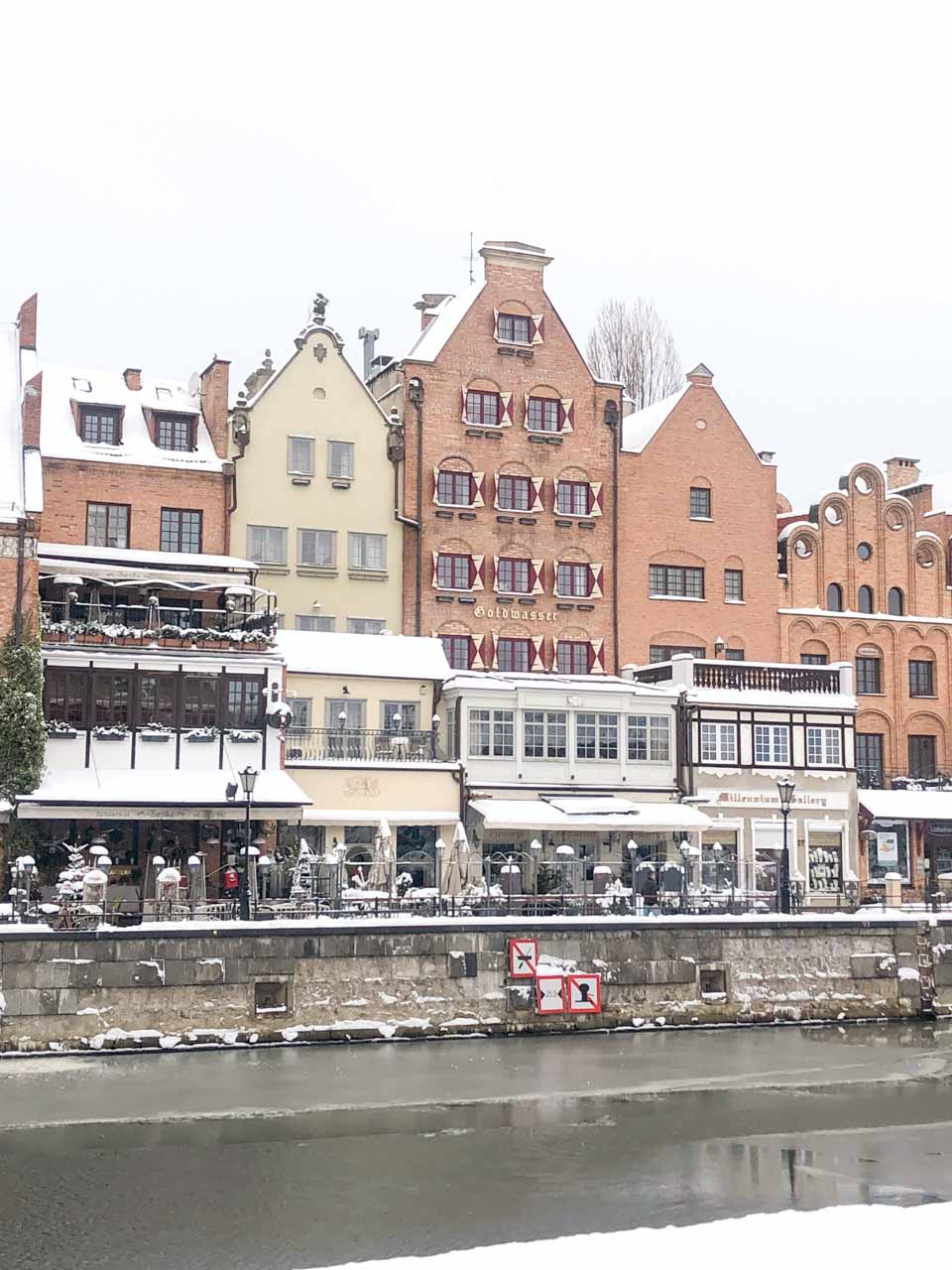 Snow-covered riverside buildings and restaurant terraces along the Motława River in Gdańsk