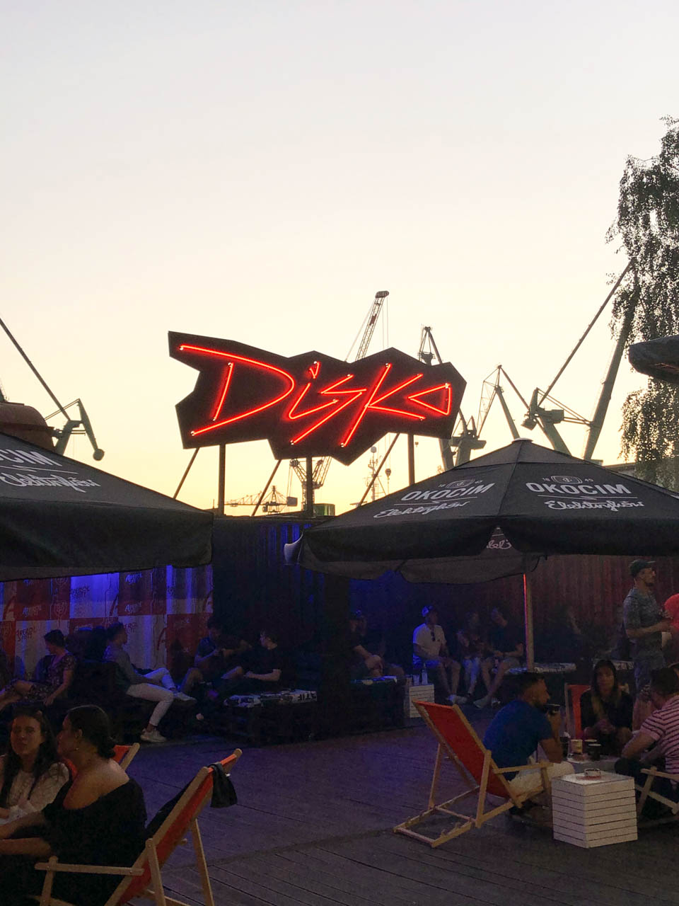 Outdoor bar on Elektryków Street in Gdańsk with a neon 'DISKO' sign and people relaxing under parasols at sunset