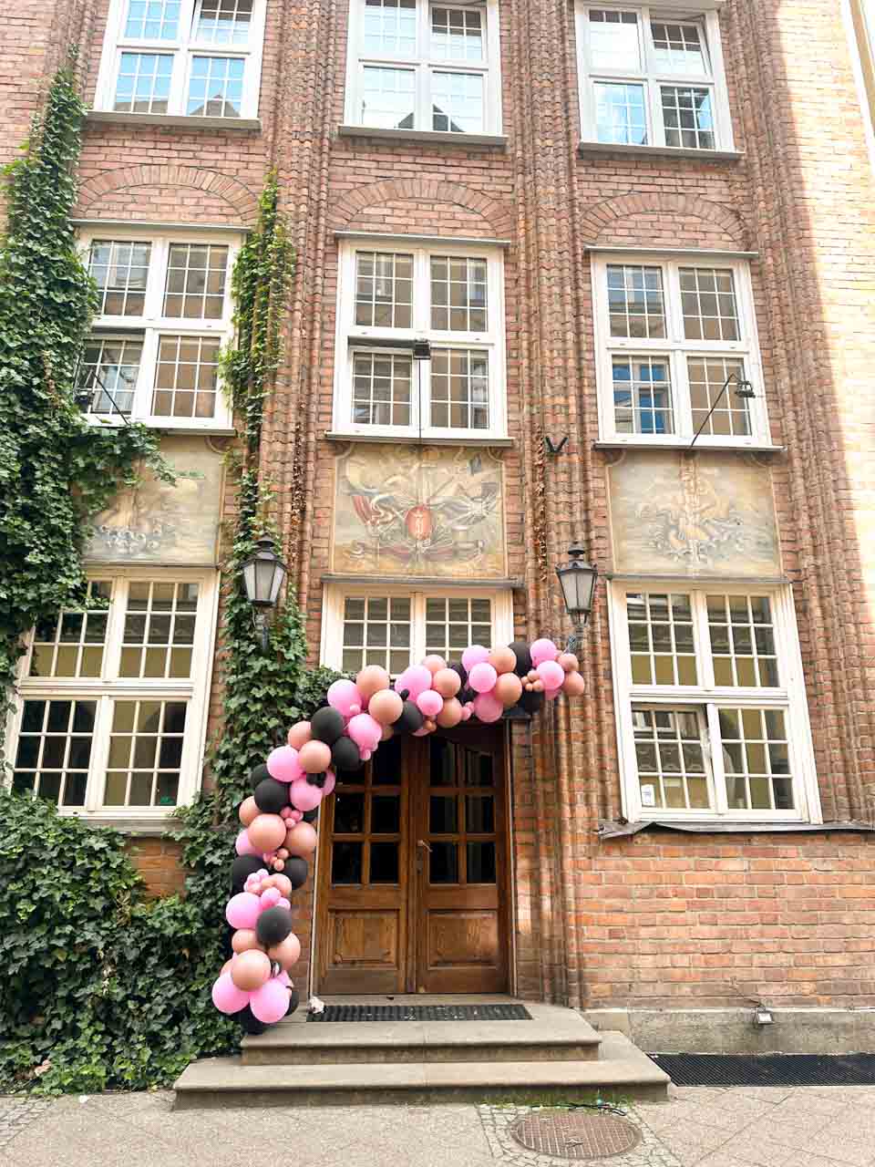 Entrance to a red brick building decorated with ivy and a pink and black balloon arch over the door