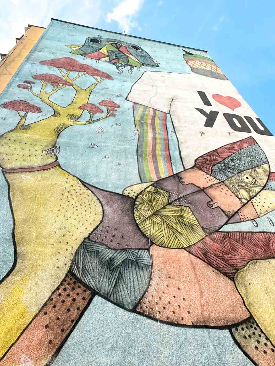 Colourful street art on a tall building featuring a bird-headed figure wearing an “I ❤️ YOU” T-shirt