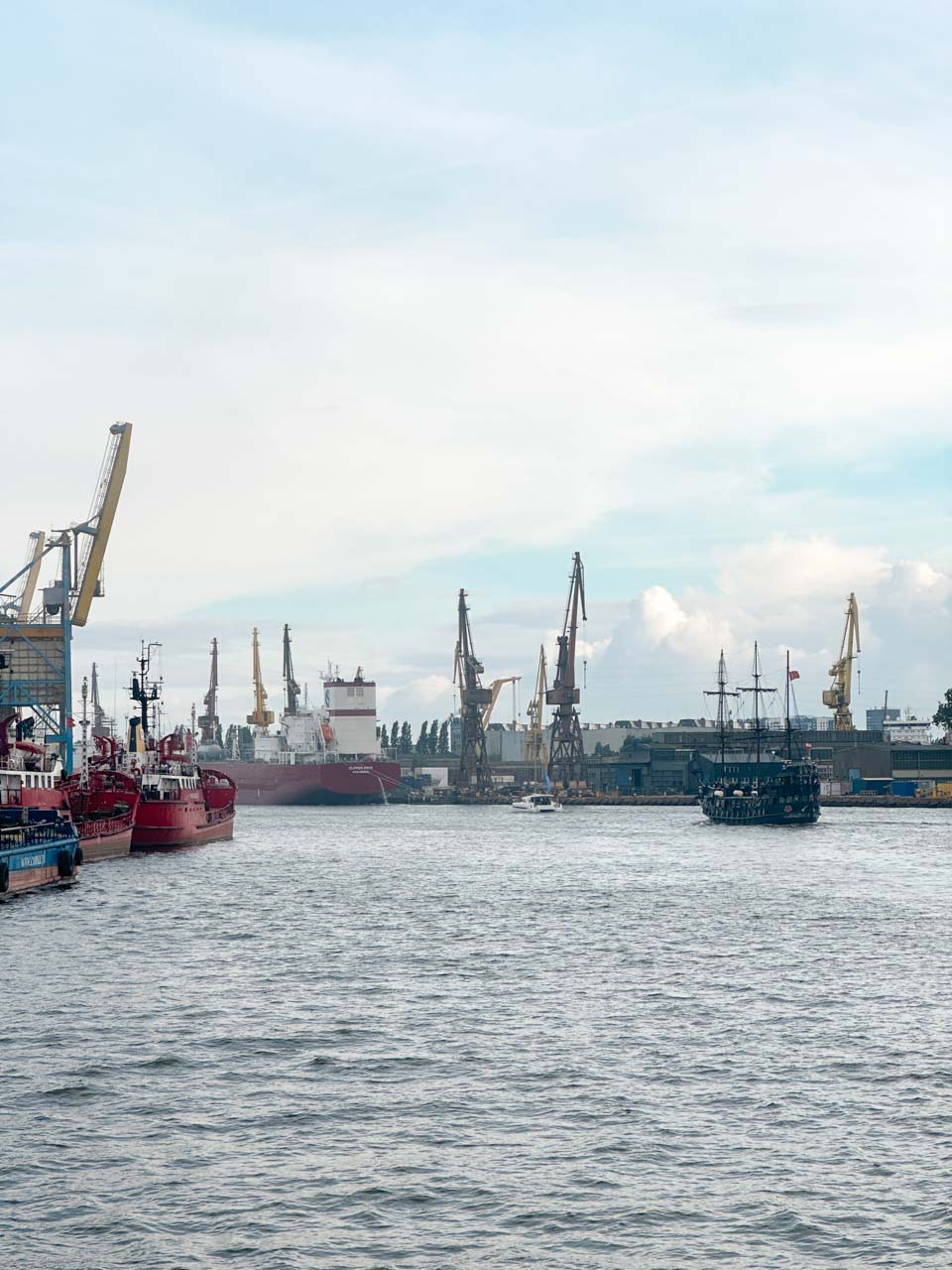 Harbour scene in Gdańsk with red ships, cargo cranes, and a black pirate-style ship on the water