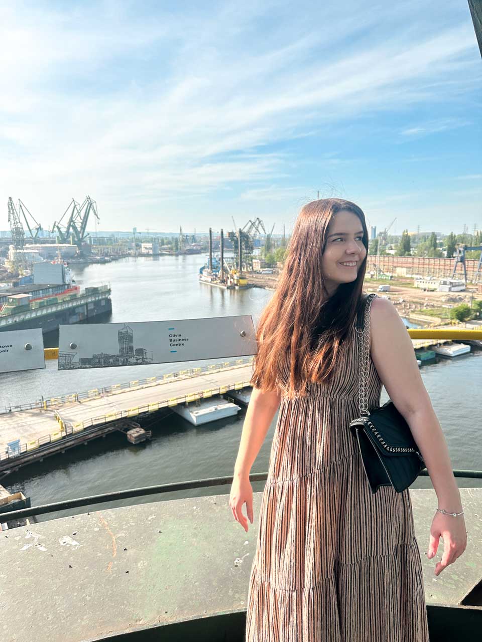 Woman in a striped dress smiling with Gdańsk shipyard cranes and the river visible behind her