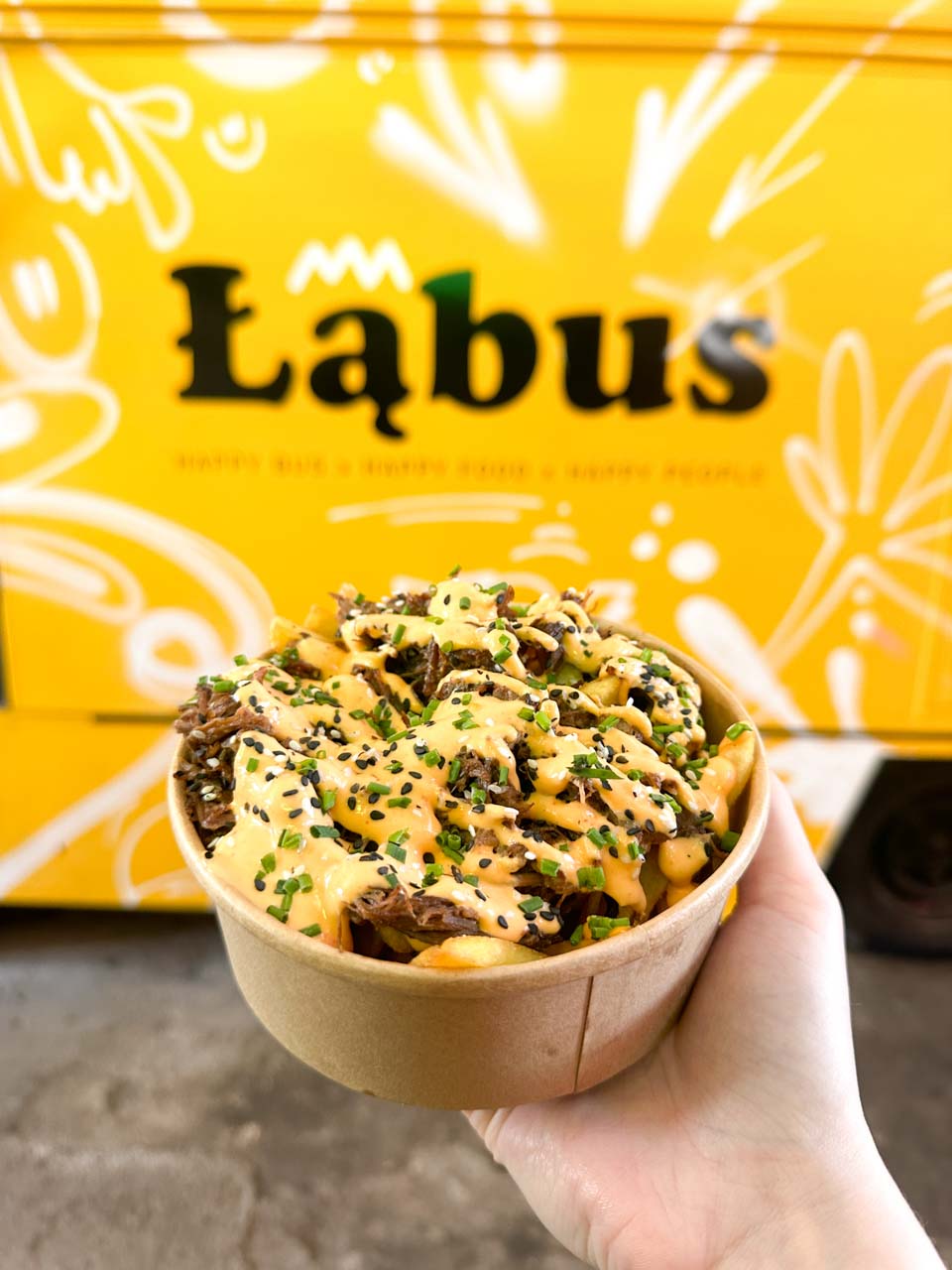 A bowl of loaded fries with shredded meat, sauce, and chives held in front of a yellow Łąbus food truck