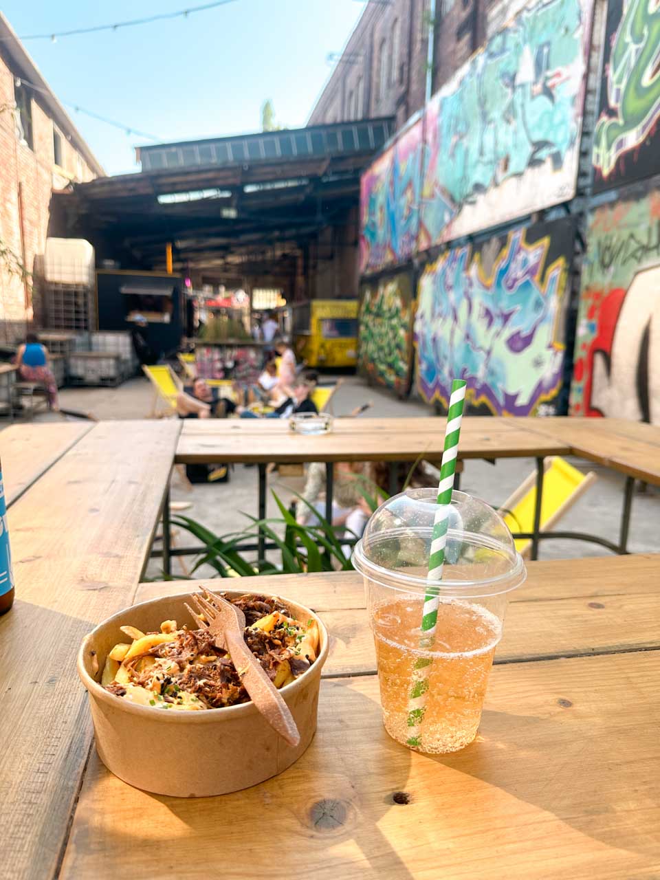 A bowl of loaded fries and a cold drink on a wooden table at the outdoor food court in 100cznia, surrounded by graffiti