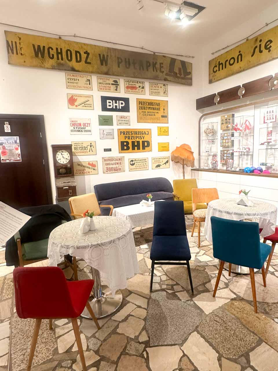 Retro-style café with colourful chairs, doily-covered tables, and vintage workplace safety signs on the wall