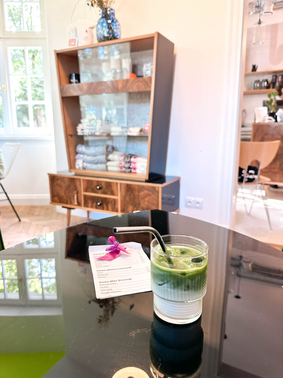 A glass of iced matcha with a metal straw on a black table, next to a printed drinks menu at Las in Gdańsk