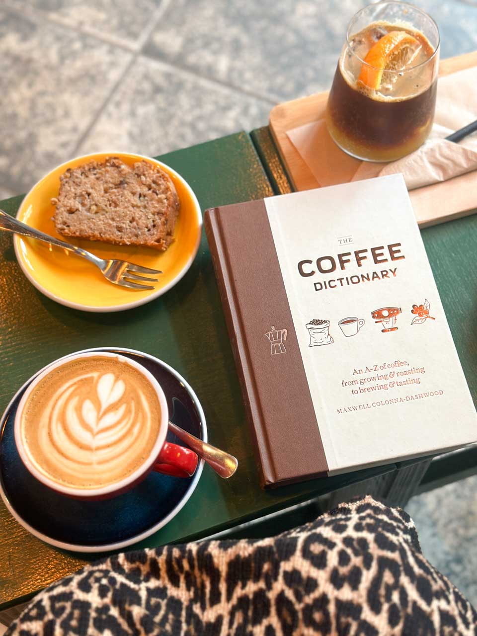A cappuccino, an orange espresso coffee, banana bread, and “The Coffee Dictionary” on a green table at Łyk Kawy in Gdańsk