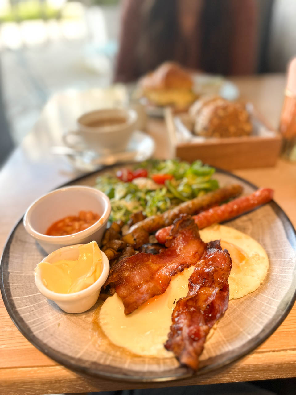 A full English breakfast with eggs, bacon, sausages, beans, mushrooms, salad, and butter on a rustic plate, served with coffee at Mielnik in Gdańsk