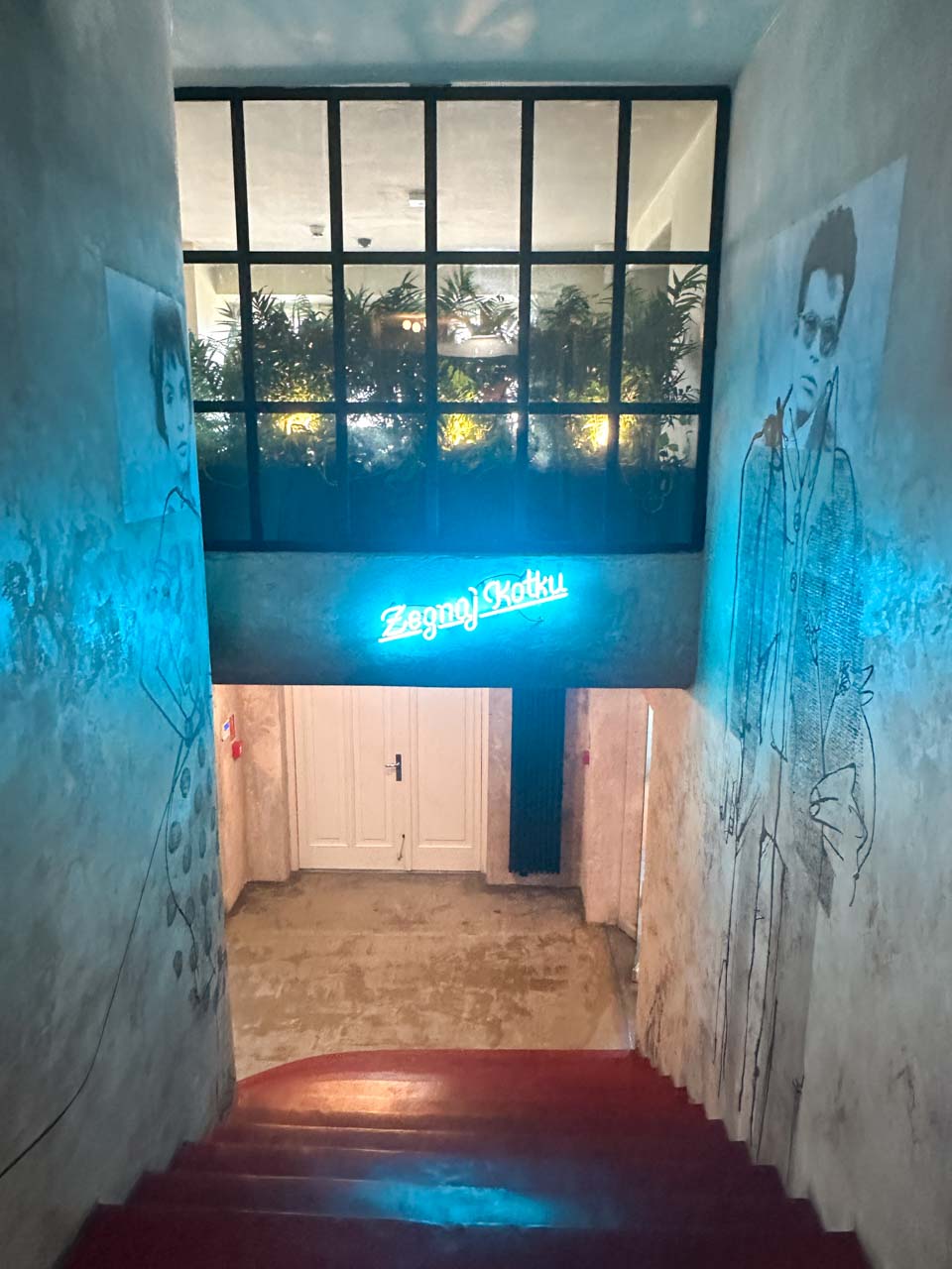 A dimly lit staircase with a blue neon sign reading “Żegnaj Kotku” and artistic portraits on the walls inside Klub KOT in Gdańsk