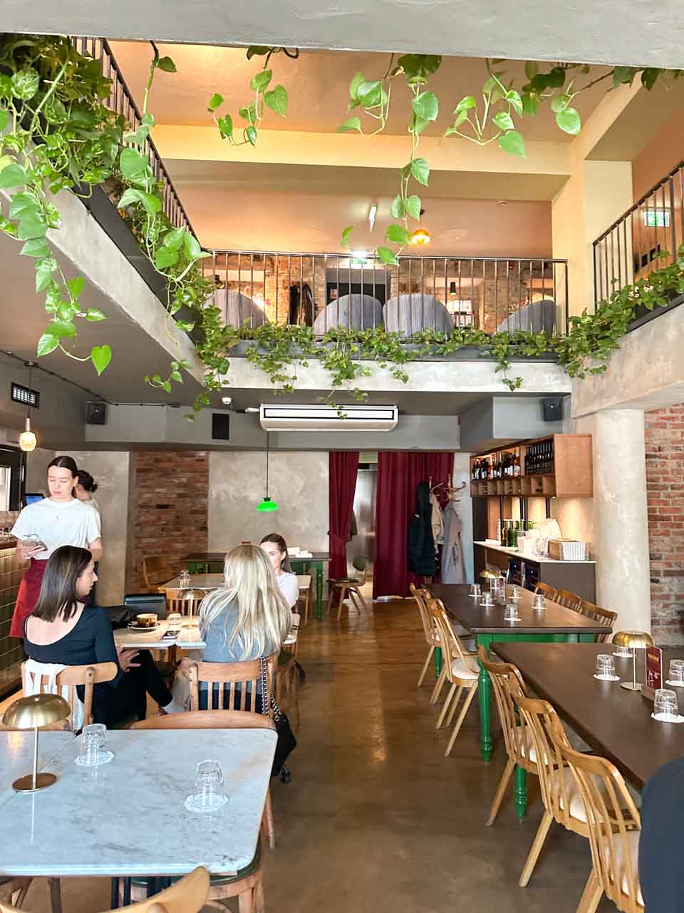 The modern interior of Masło Maślane in Gdańsk with two levels, hanging plants, and people enjoying breakfast at one of the marble tables