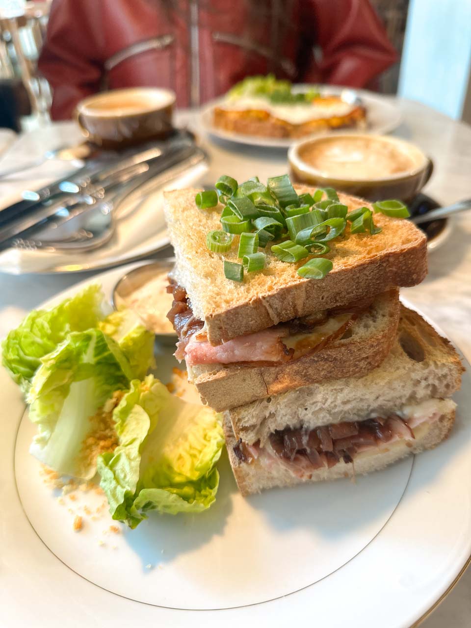 Close-up of a toasted sandwich layered with bacon and onions, garnished with chopped spring onions and romaine lettuce at Masło Maślane in Gdańsk