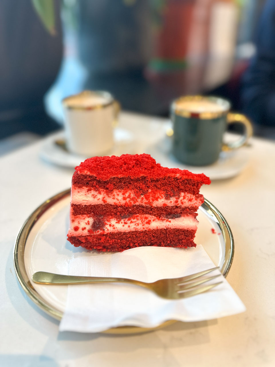 Slice of red velvet cake with cream cheese frosting on a gold-rimmed plate next to elegant coffee cups at Kuchy i Qruchy in Gdańsk