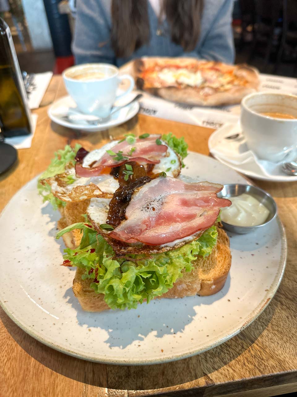 Open-faced toast with lettuce, bacon, fried egg, and caramelised onions, served with coffee and a pizza in the background at Aioli in Gdańsk