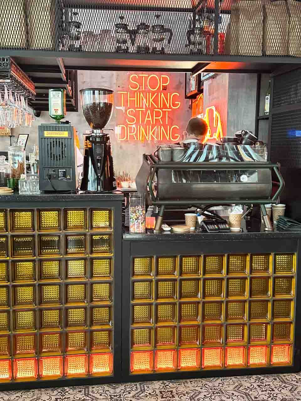 The bar area at Przystanek Garnizon in Gdańsk with yellow glass blocks, a neon sign reading "Stop Thinking Start Drinking," and coffee equipment