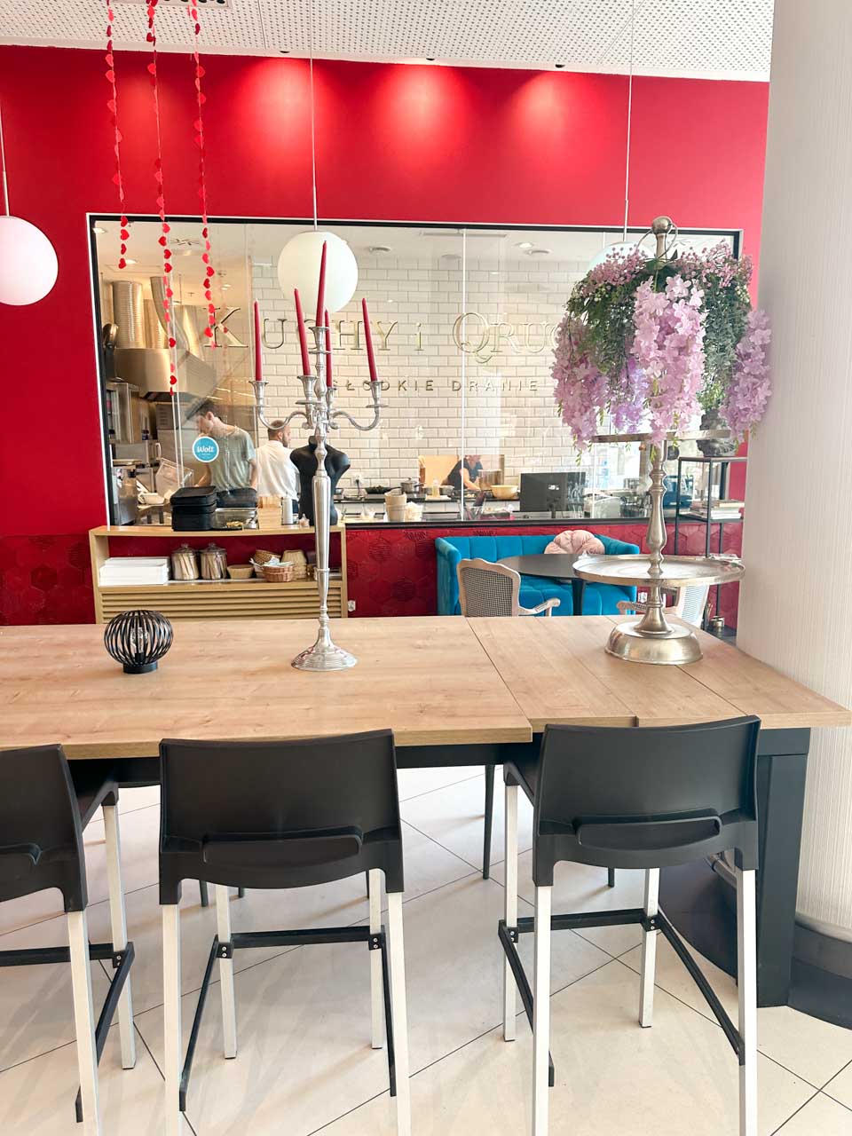 The bright interior of Kuchy i Qruchy in Gdańsk with a long wooden table, red accent wall, and a glass window into the kitchen