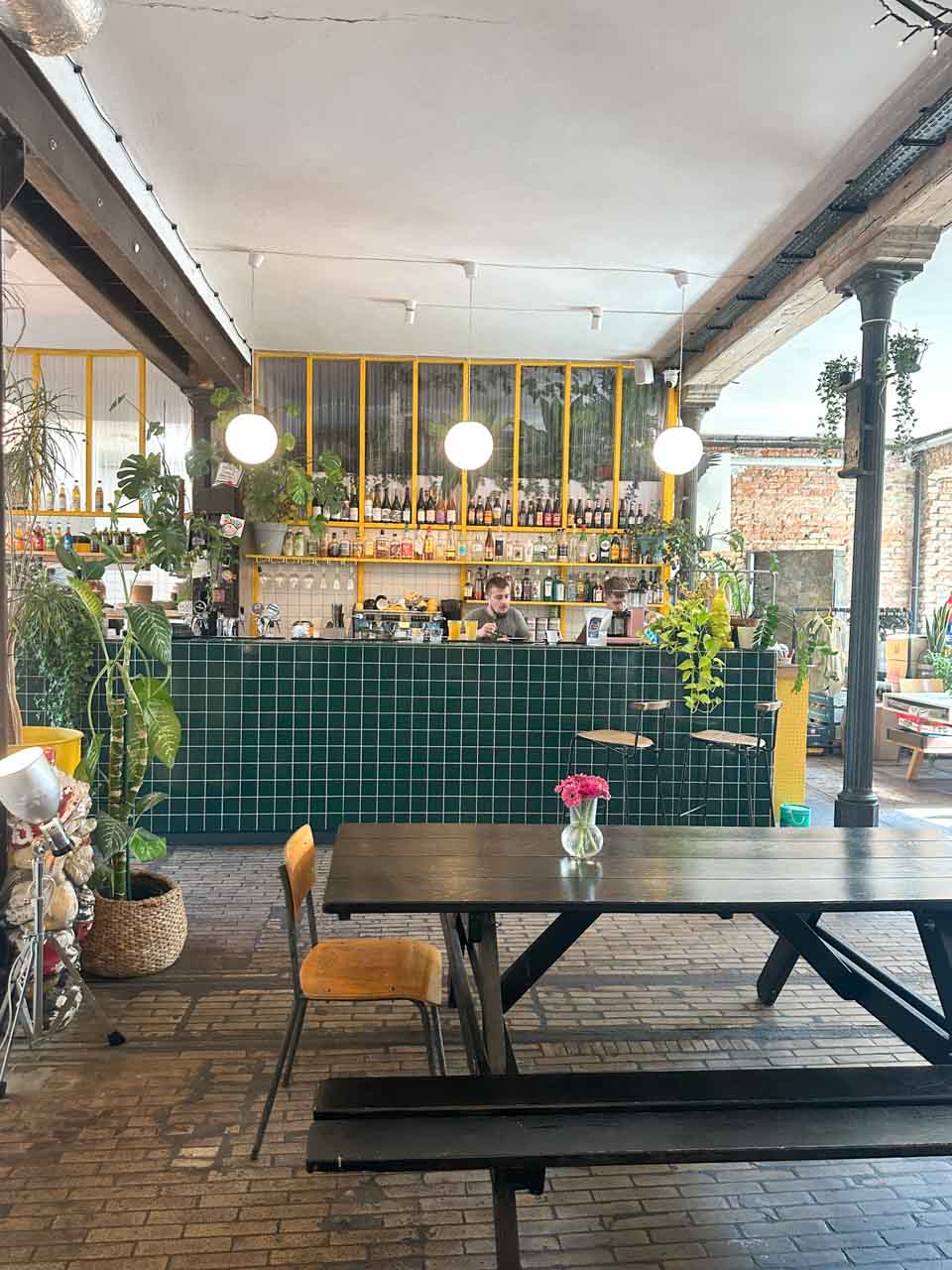 The interior of Łąka Bar in Gdańsk with green tiles, yellow accents, a long wooden table, and a bar stocked with bottles and plants