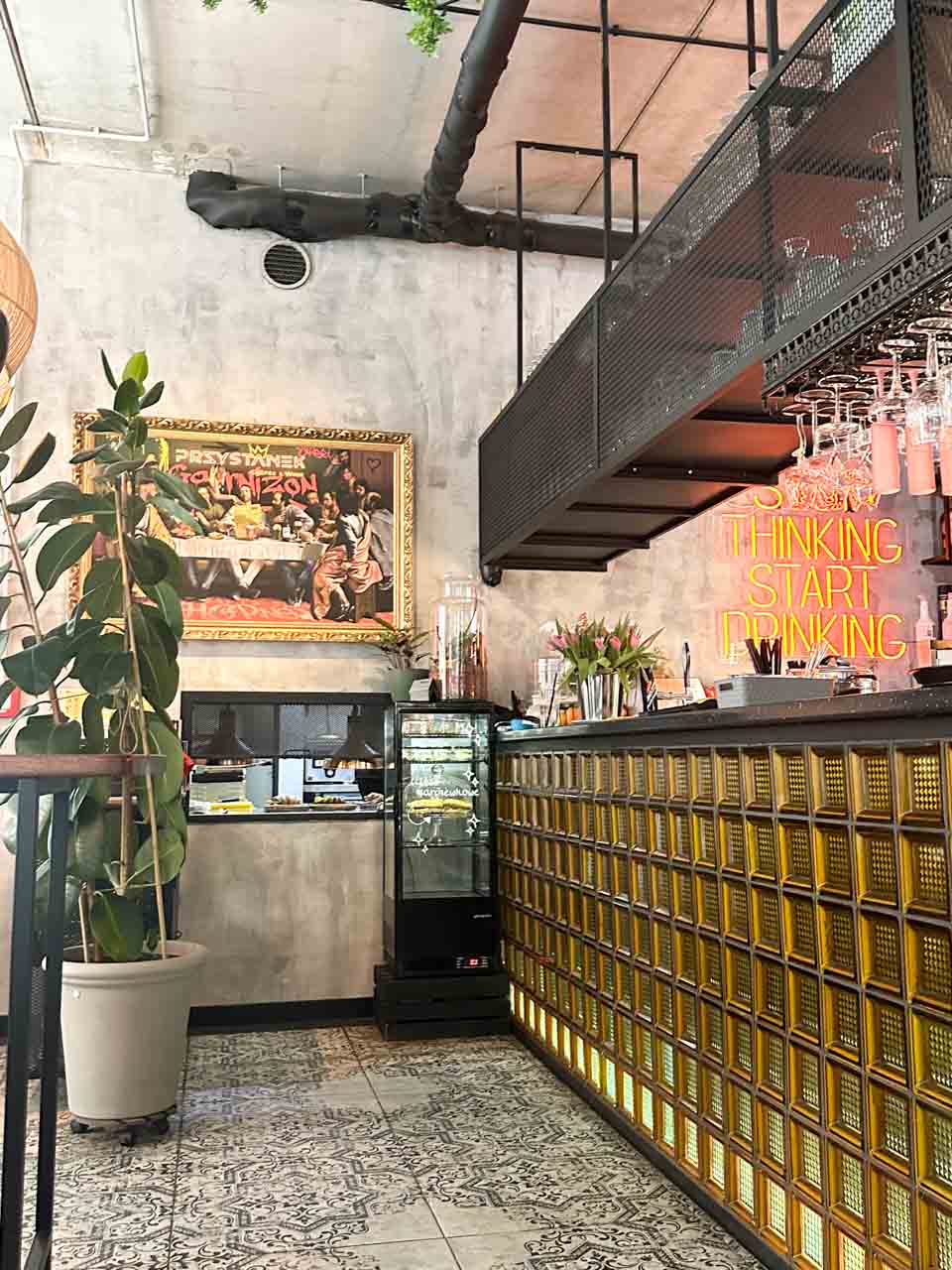 The interior of Przystanek Garnizon in Gdańsk with a tiled bar, potted plants, a neon wall sign, and a framed painting with text