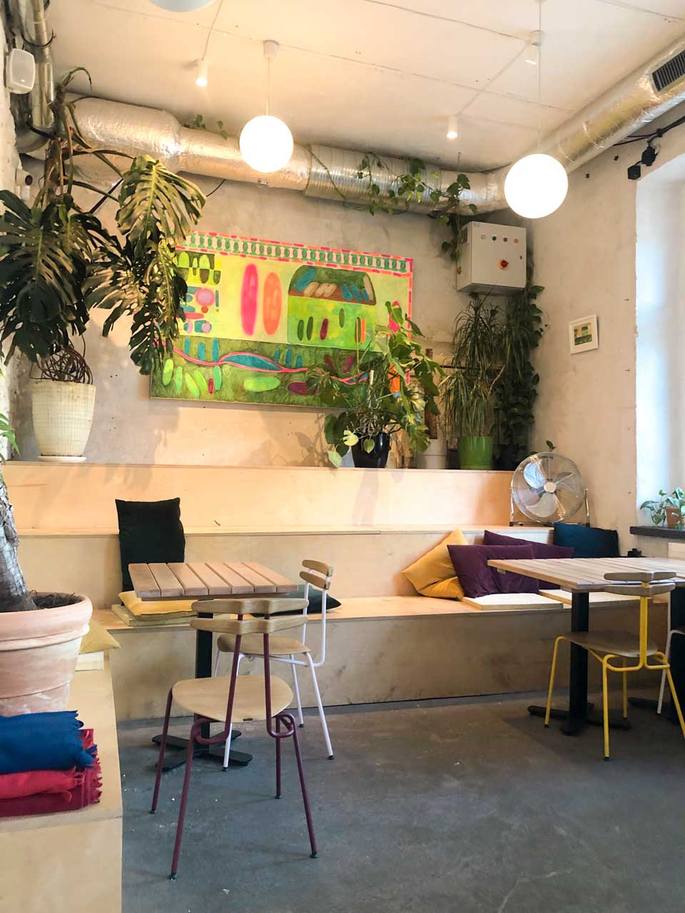A cosy corner at Łąka Bar in Gdańsk with colourful chairs, hanging plants, and a bright abstract painting on a raw concrete wall