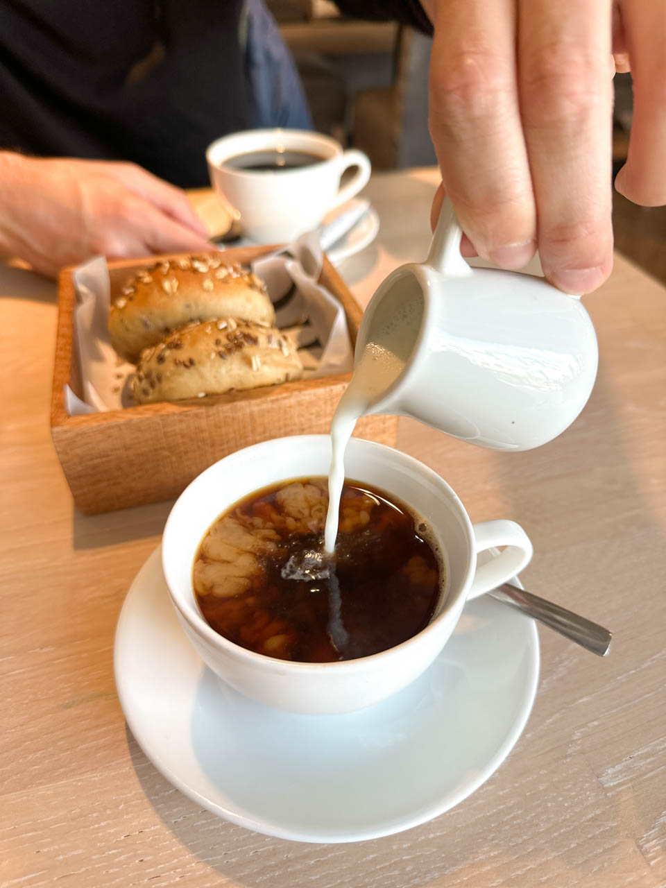 Milk being poured into a black coffee, with a basket of seeded bread rolls and a cup of coffee in the background