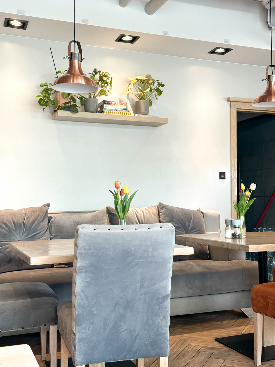 Cosy interior of Mielnik in Gdańsk with velvet chairs, wooden tables, and tulips in vases, with books and plants on a shelf above the seating area