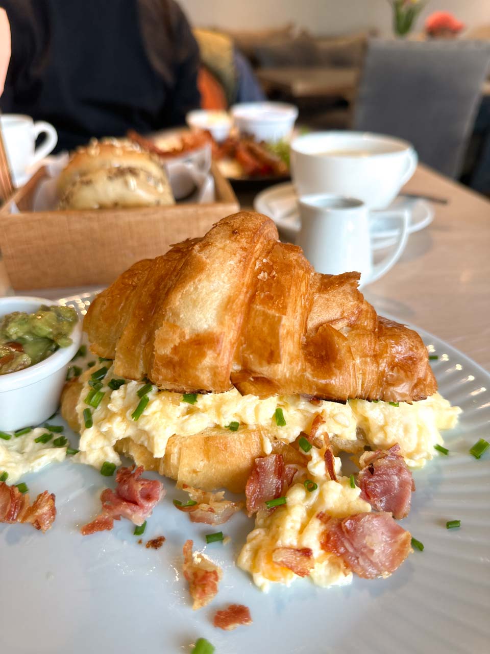 Close-up of a buttery croissant sandwich with scrambled eggs and bacon, with coffee and bread rolls in the background