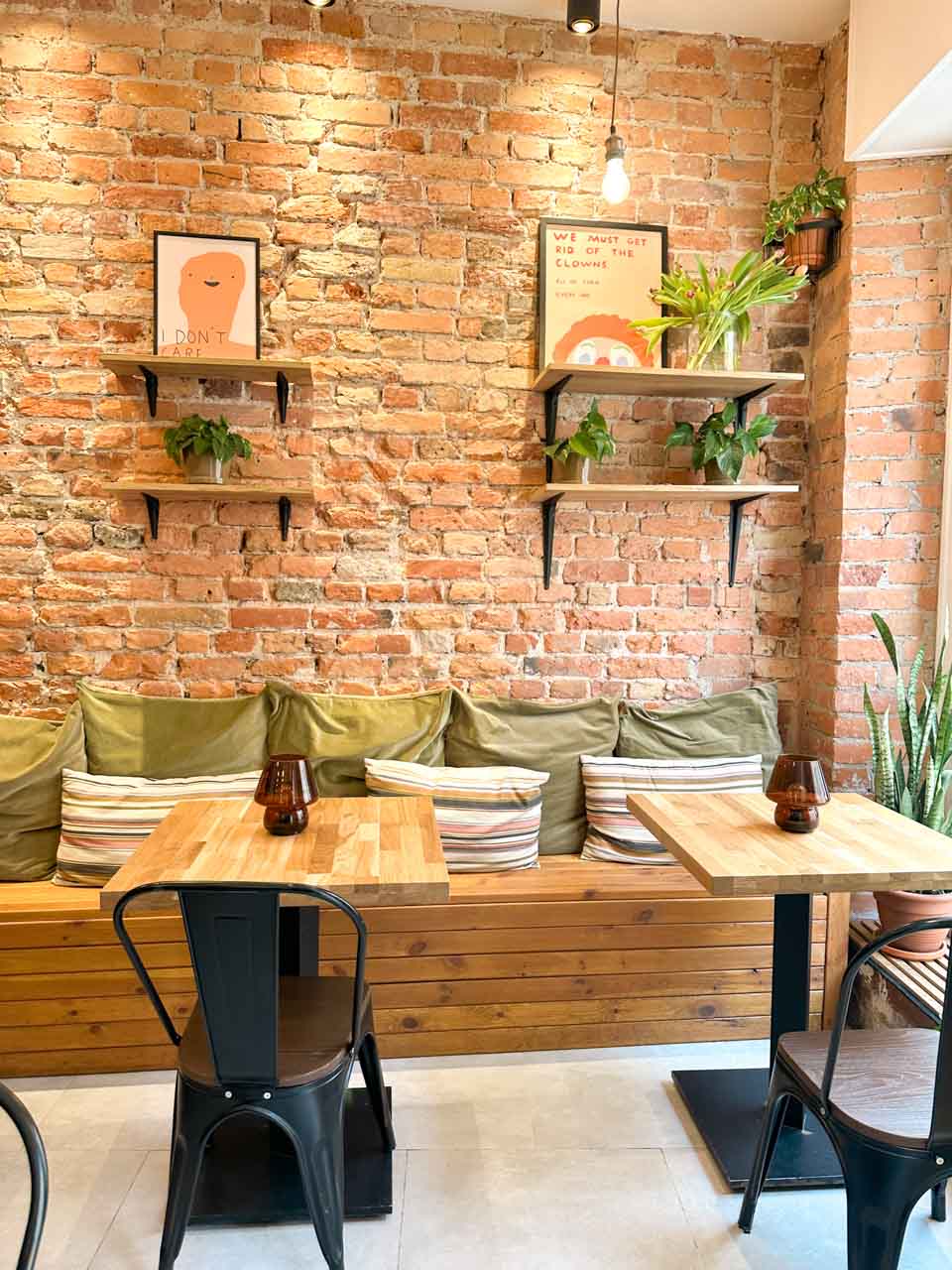Cosy brick-wall café corner with green cushions, striped pillows, plants, and framed quirky art prints on the shelves