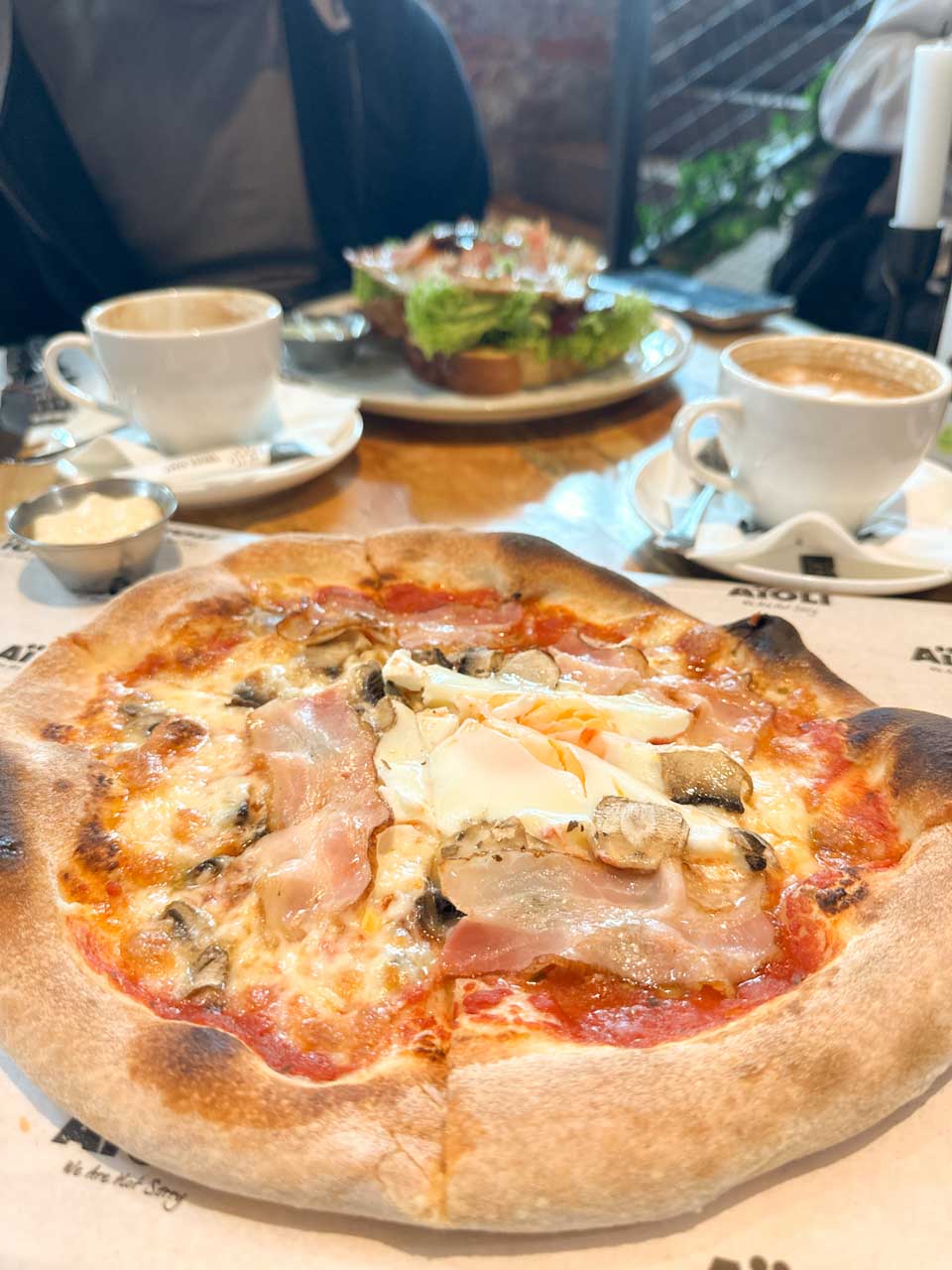 Thin-crust pizza topped with ham, mushrooms, and cheese, served with coffee and an open toast sandwich in the background at Aioli in Gdańsk