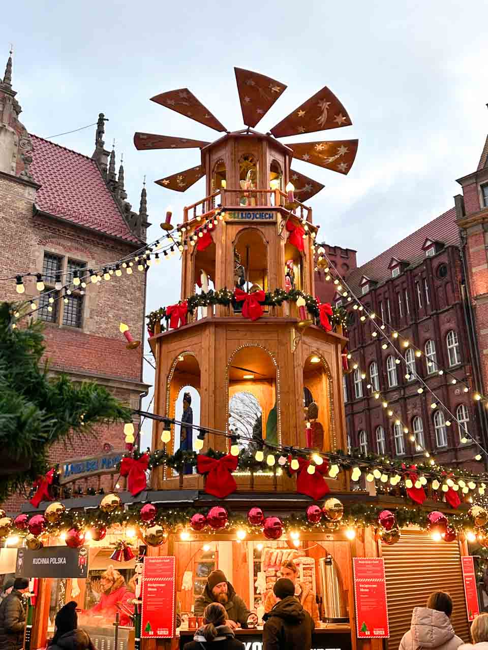 A large wooden Christmas pyramid with figurines and spinning blades, surrounded by festive lights and visitors