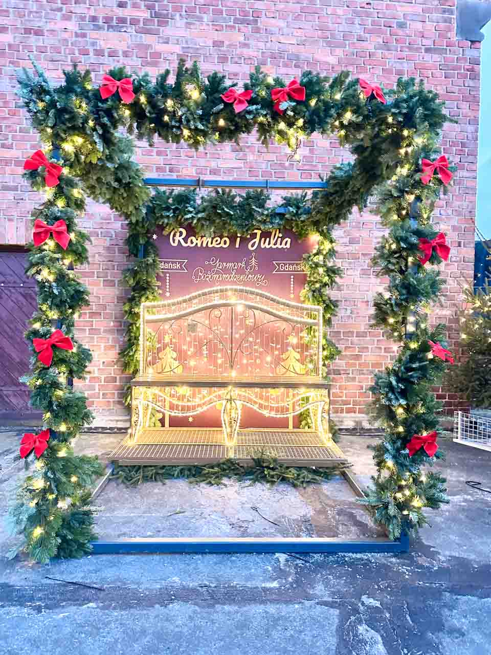 A romantic Christmas photo spot featuring a light-up bench under a garland arch with red bows, marked “Romeo i Julia.”