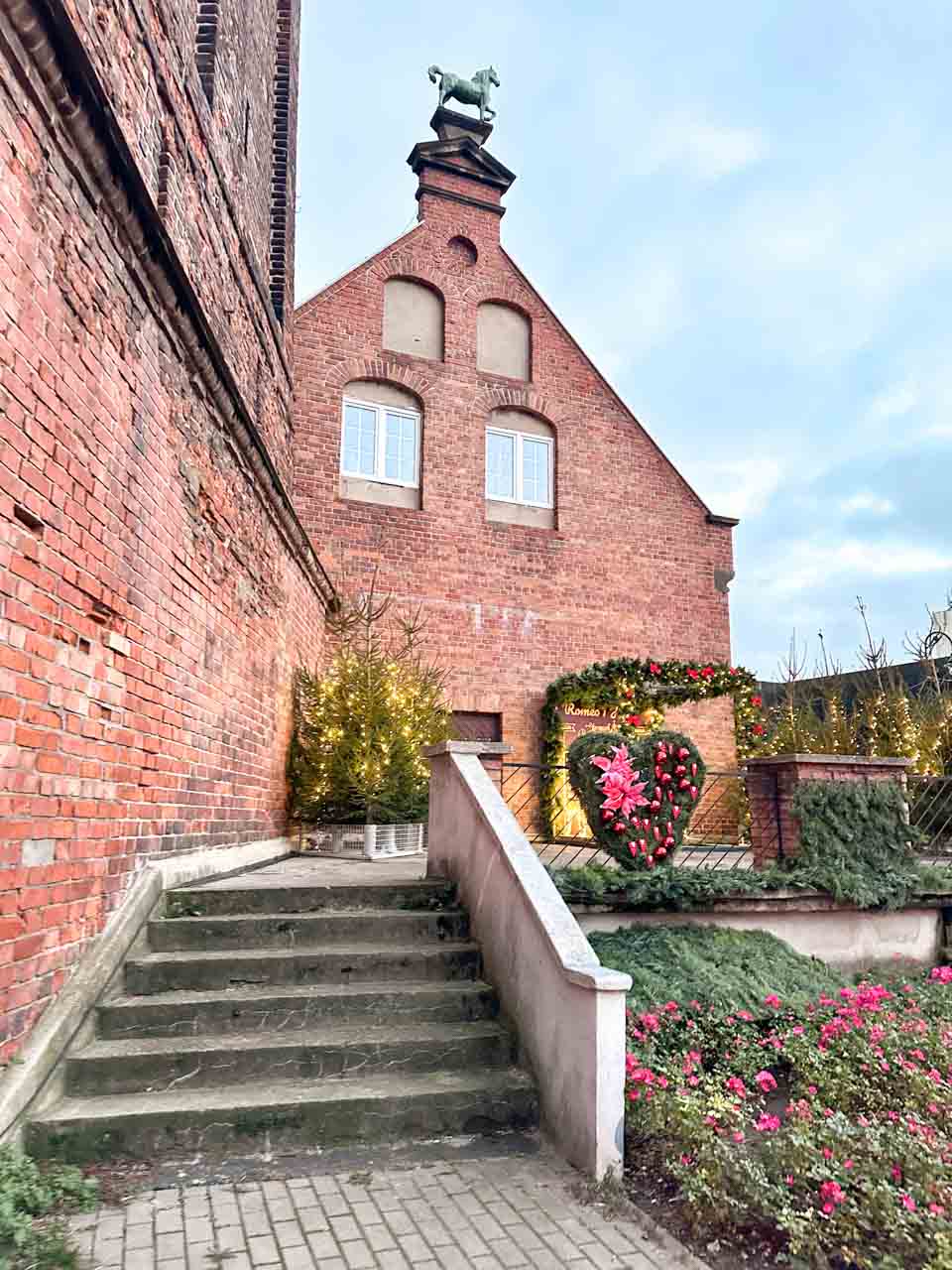 A side view of a red-brick building topped with a horse statue, with Christmas trees and a heart-shaped floral wreath nearby