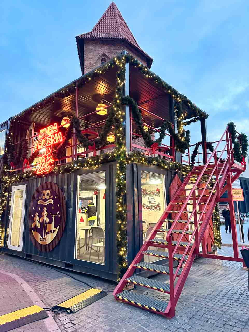 A festive food stall decorated with fairy lights and garlands, with a neon chicken sign above