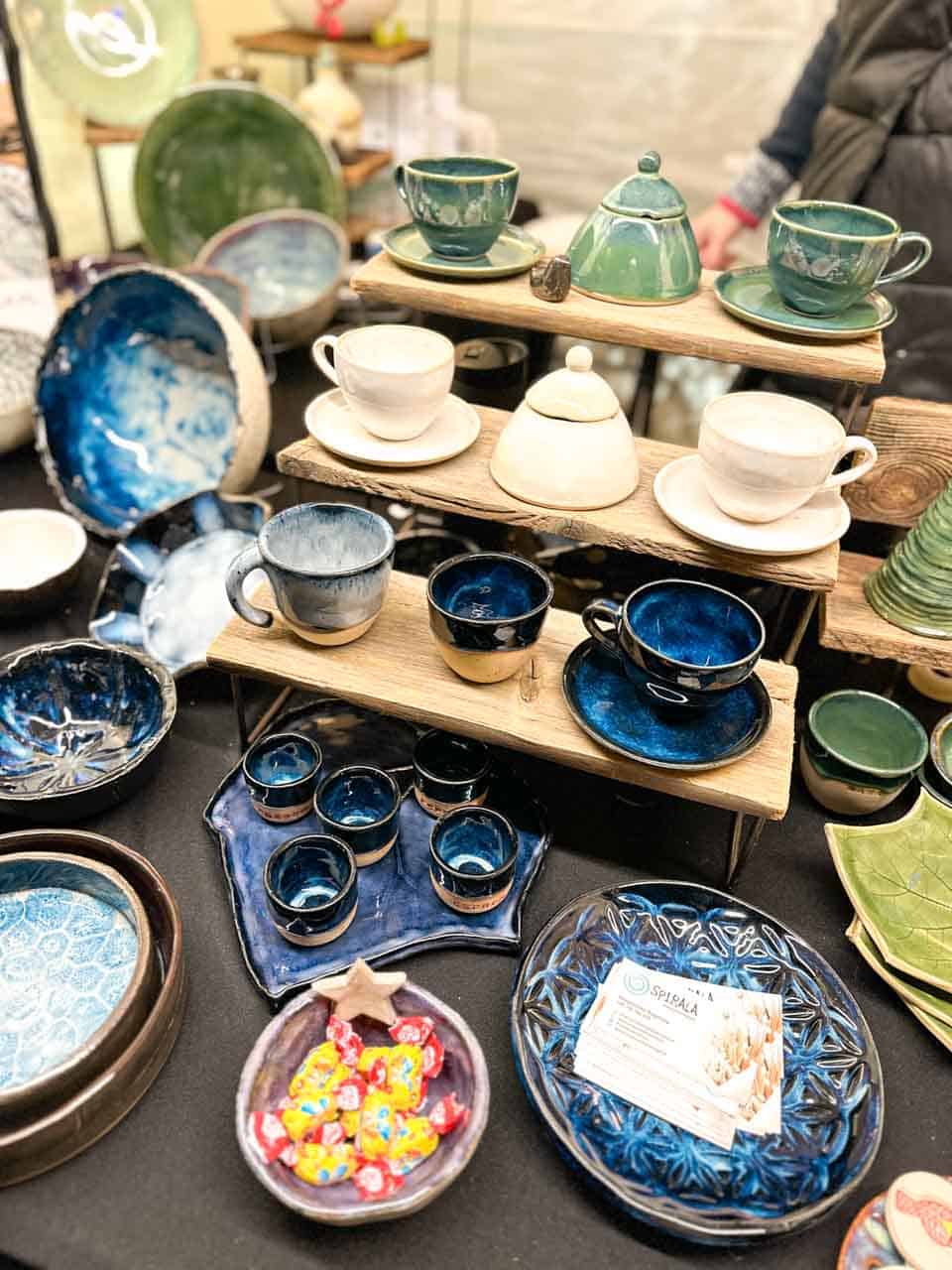 Handmade ceramic cups, bowls, and plates in green, cream, and deep blue displayed on a wooden stand at the Krak&oacute;w Christmas market