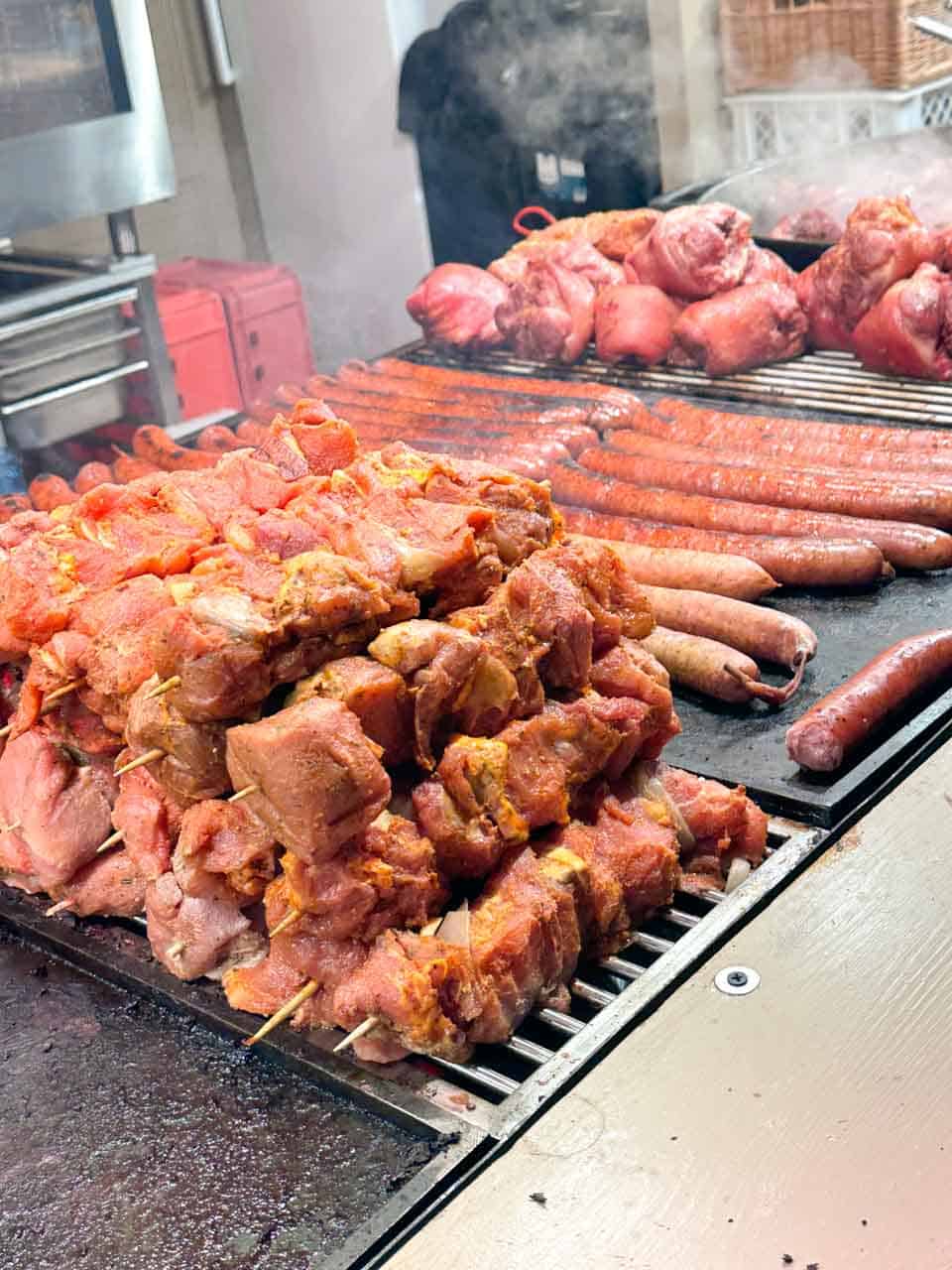 Close-up of a sizzling grill stacked with marinated meat skewers, sausages, and smoked pork knuckles at the Krak&oacute;w Christmas market